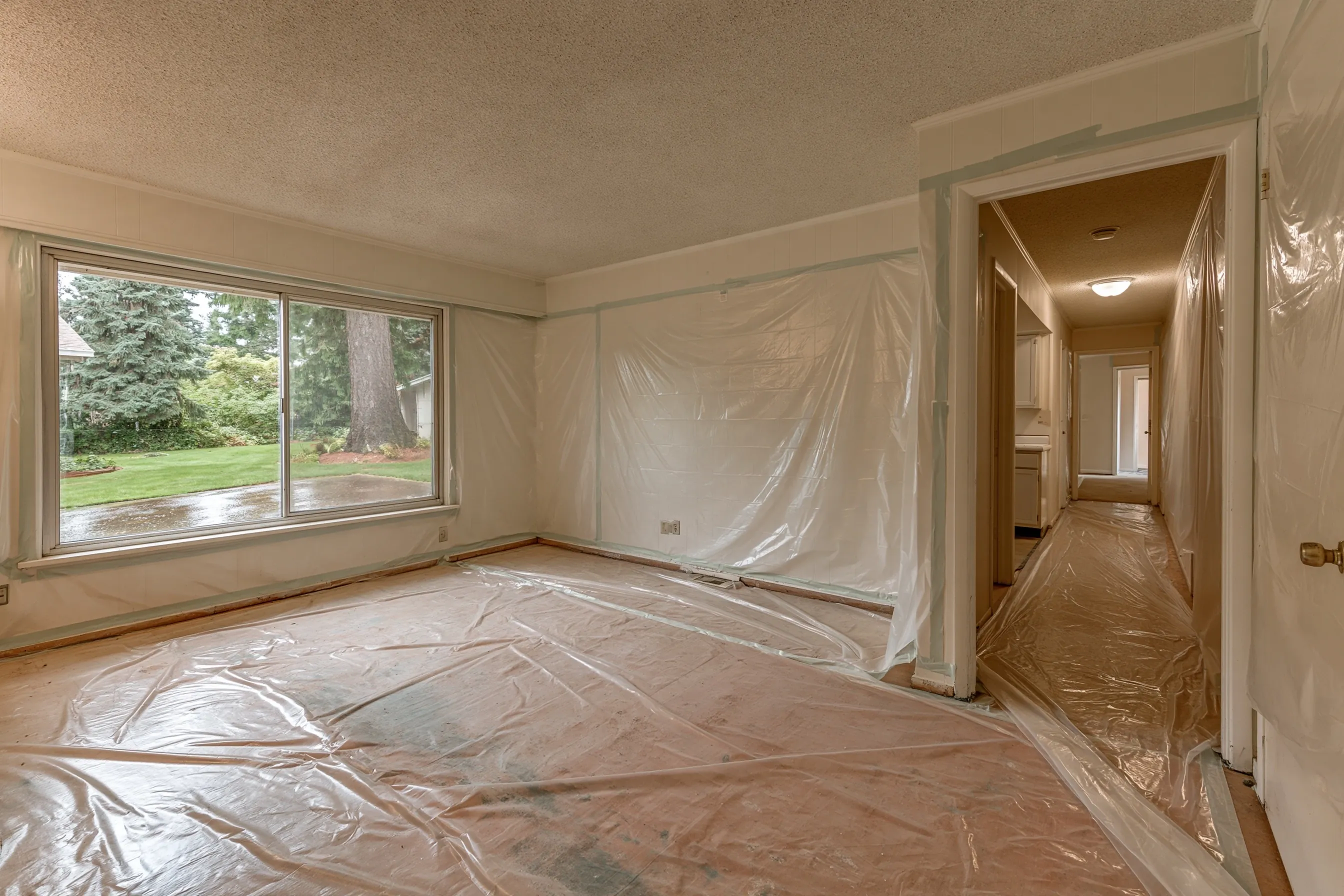 Empty one-story 1970s living room and hallway with popcorn ceilings, bare floors and a roll of plastic sheeting